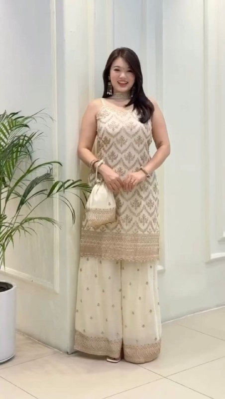 Woman in a traditional outfit standing indoors with a plant and white curtain in the background