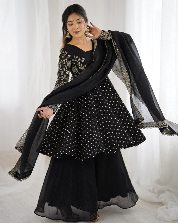 Woman wearing a black traditional outfit with a sheer dupatta against a white curtain background