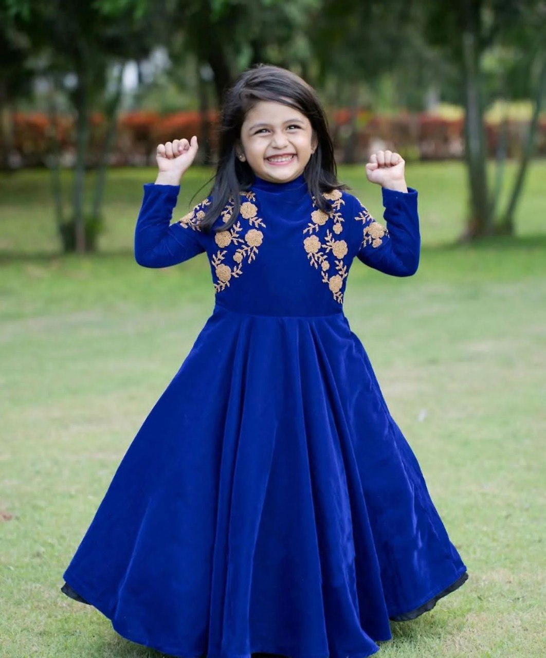 Young girl in a blue dress with gold embroidery standing outdoors on grass.