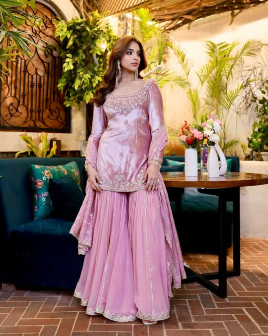 Woman in a pink traditional outfit standing in an indoor setting with plants and decor.