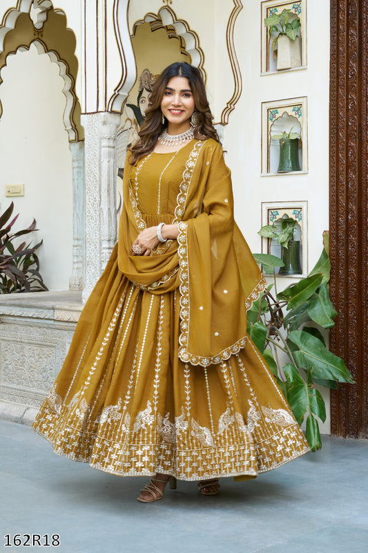 Woman in a mustard yellow traditional outfit with intricate patterns standing in an ornate interior setting.