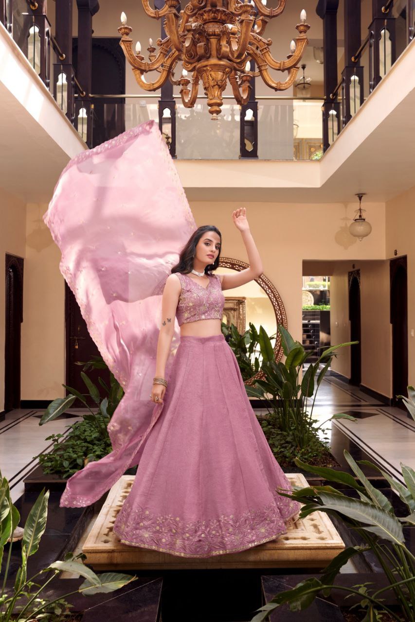 Woman in a pink traditional outfit standing in a decorated indoor setting with plants and a chandelier.