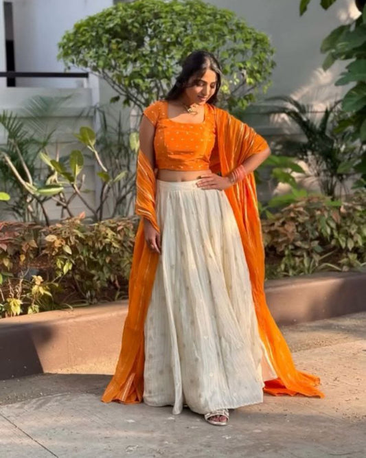 Woman in an orange and white traditional outfit standing outdoors with greenery in the background