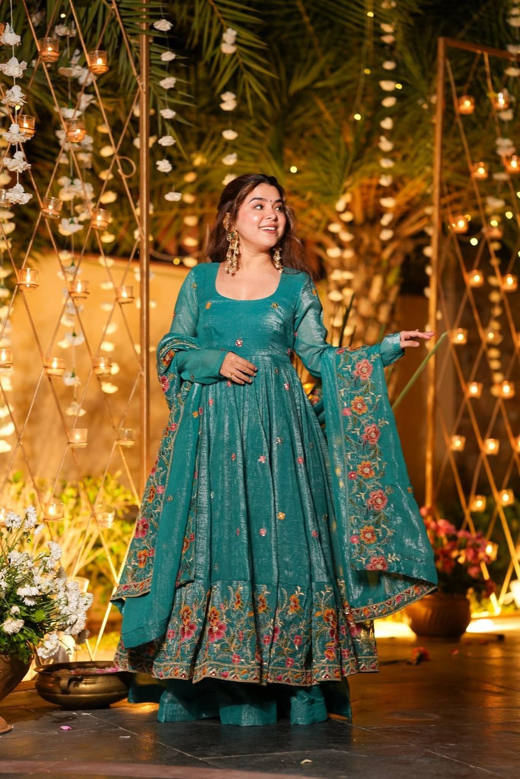 Woman in a teal traditional outfit with floral patterns standing in front of decorative lights and plants.