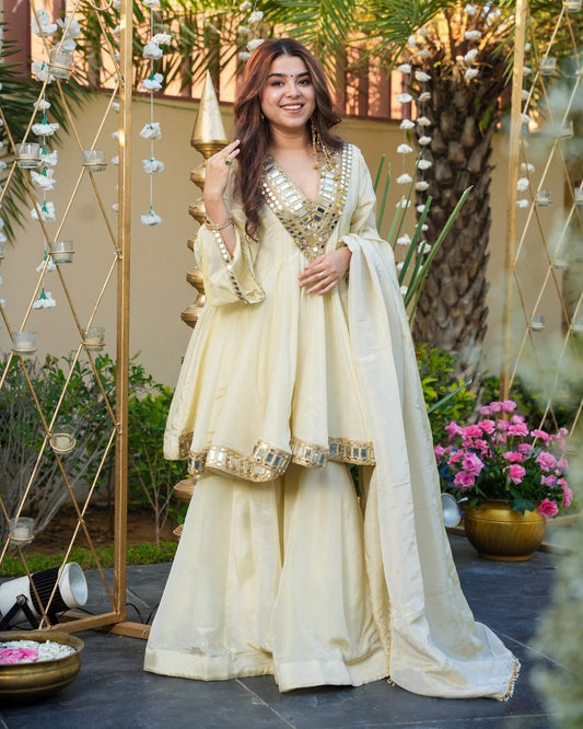 Woman in a traditional cream-colored outfit standing outdoors with decorative elements and plants.