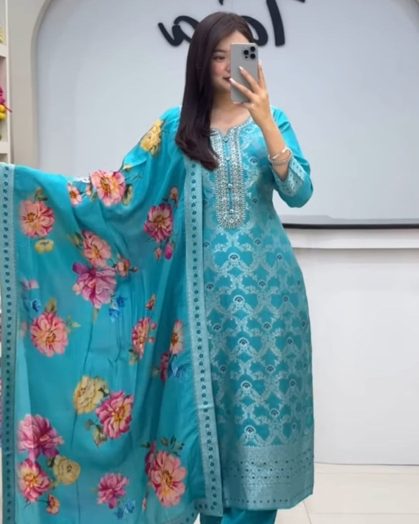 Woman wearing a turquoise embroidered outfit with floral patterns, standing in front of a white wall with a logo.