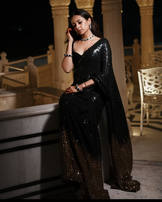 Woman in a black sequined saree sitting on a stone ledge at night.