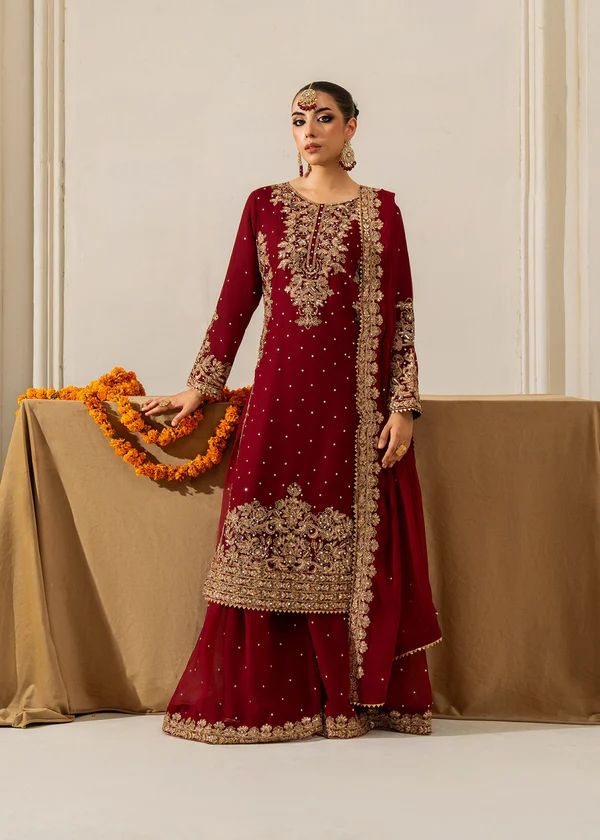 Woman in a red and gold embroidered traditional outfit holding a garland against a beige wall.