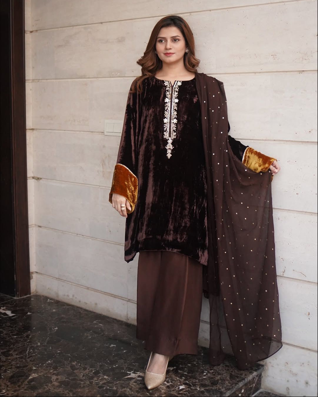 Woman wearing a brown traditional outfit with a matching dupatta against a light-colored wall.
