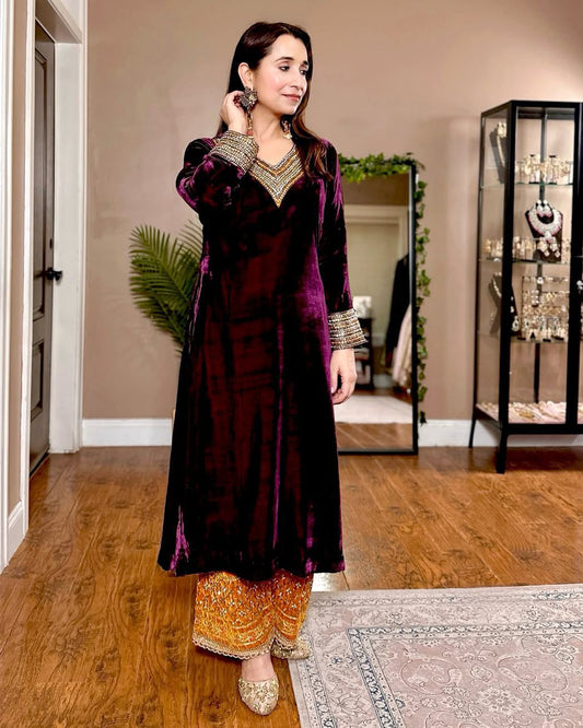 Woman in a dark purple traditional outfit with gold patterns in a room with wooden flooring and decorative items.