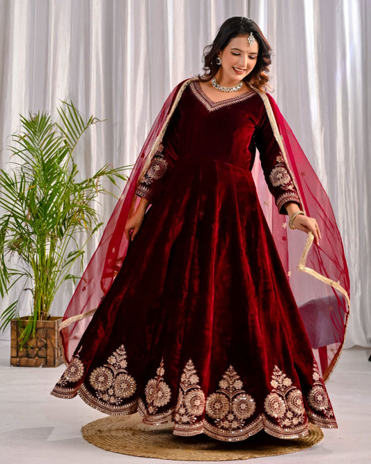 Woman in a maroon embroidered traditional outfit with a red dupatta against a white curtain background.