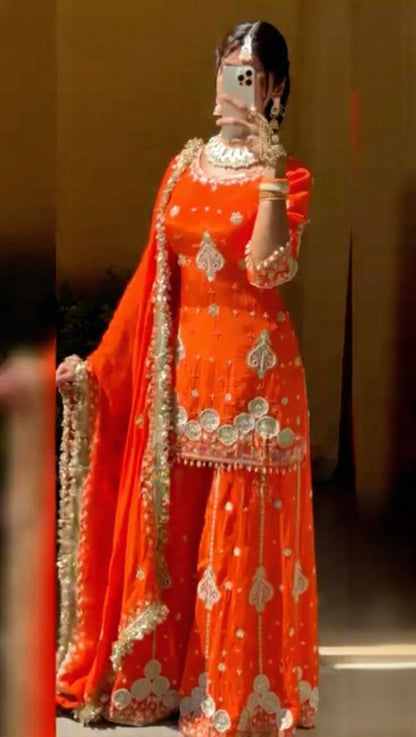 Person wearing an orange traditional outfit with intricate designs, standing against a neutral background.