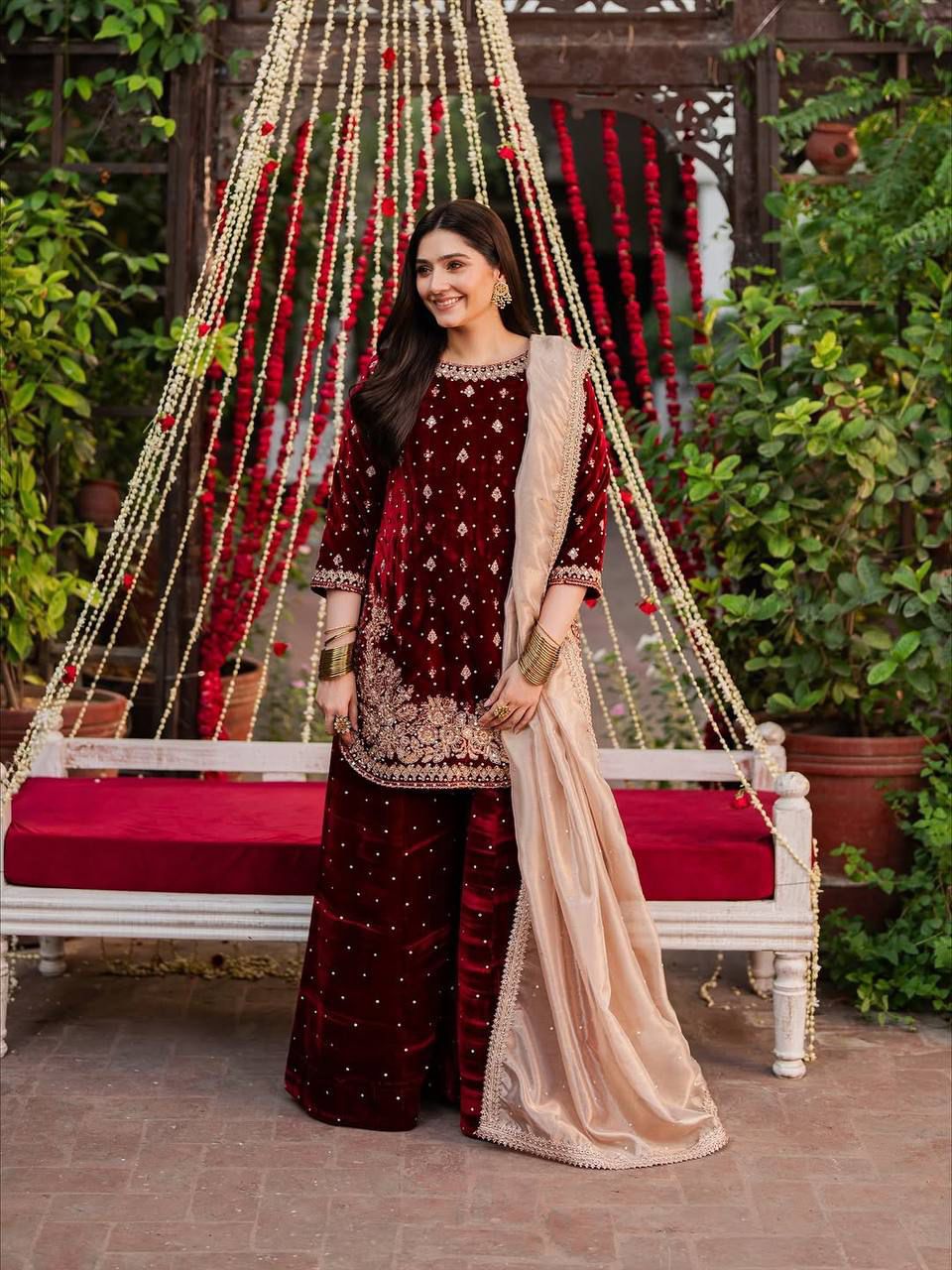 Woman in traditional maroon and beige outfit standing in front of decorative hanging structure with plants.
