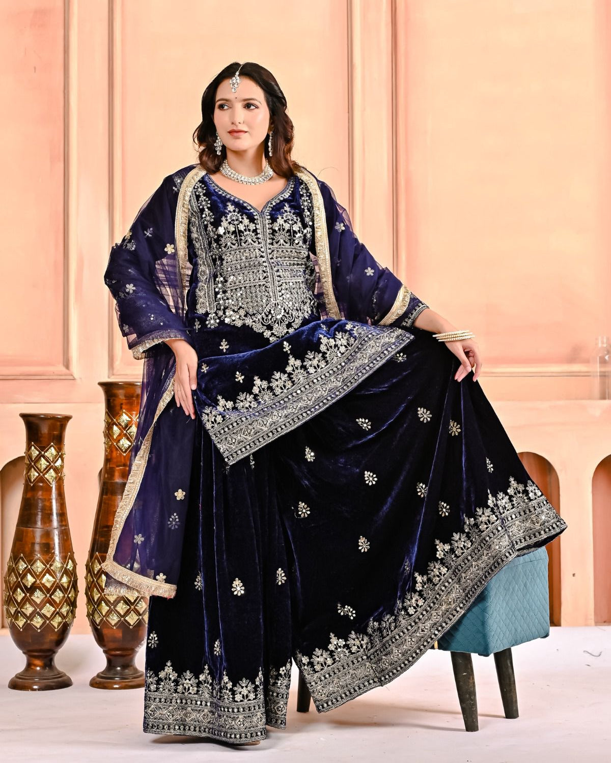 Woman in a traditional embroidered outfit sitting on a chair against a pink wall.