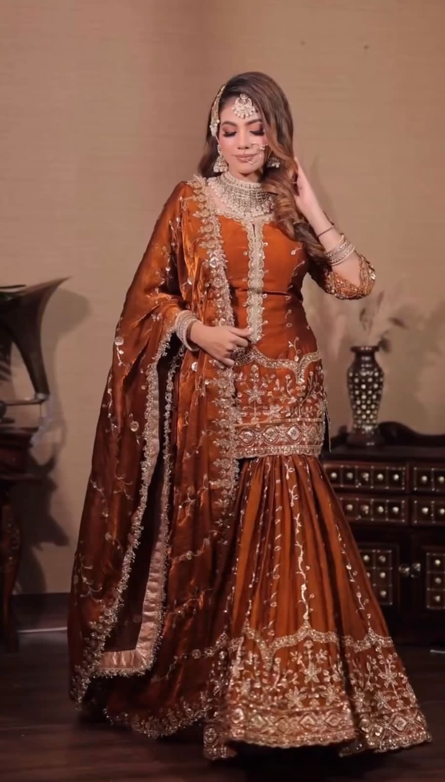 Woman in a traditional brown and gold embroidered outfit with jewelry, standing indoors.