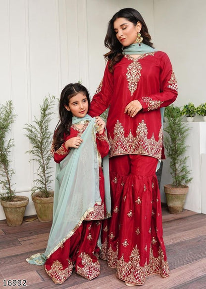Two women in red embroidered traditional outfits standing indoors with plants in the background.