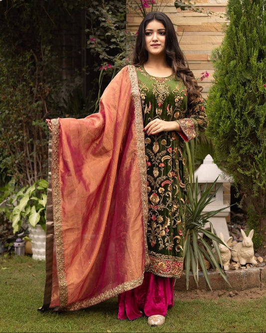 Woman in traditional green and pink embroidered outfit standing outdoors with plants and a stone lantern in the background.