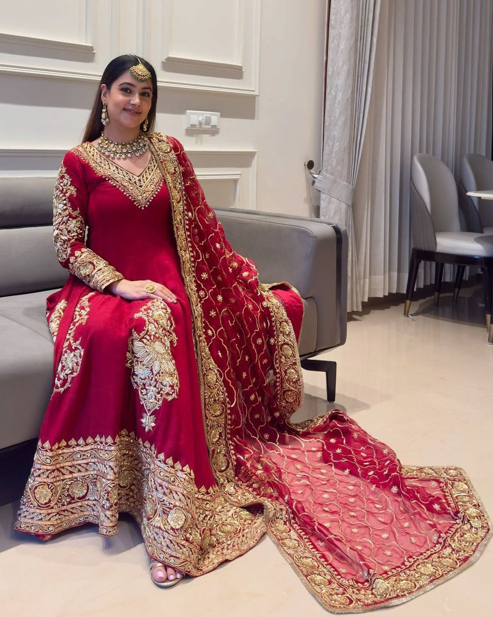 Woman in a red and gold traditional outfit sitting on a couch in a modern room.